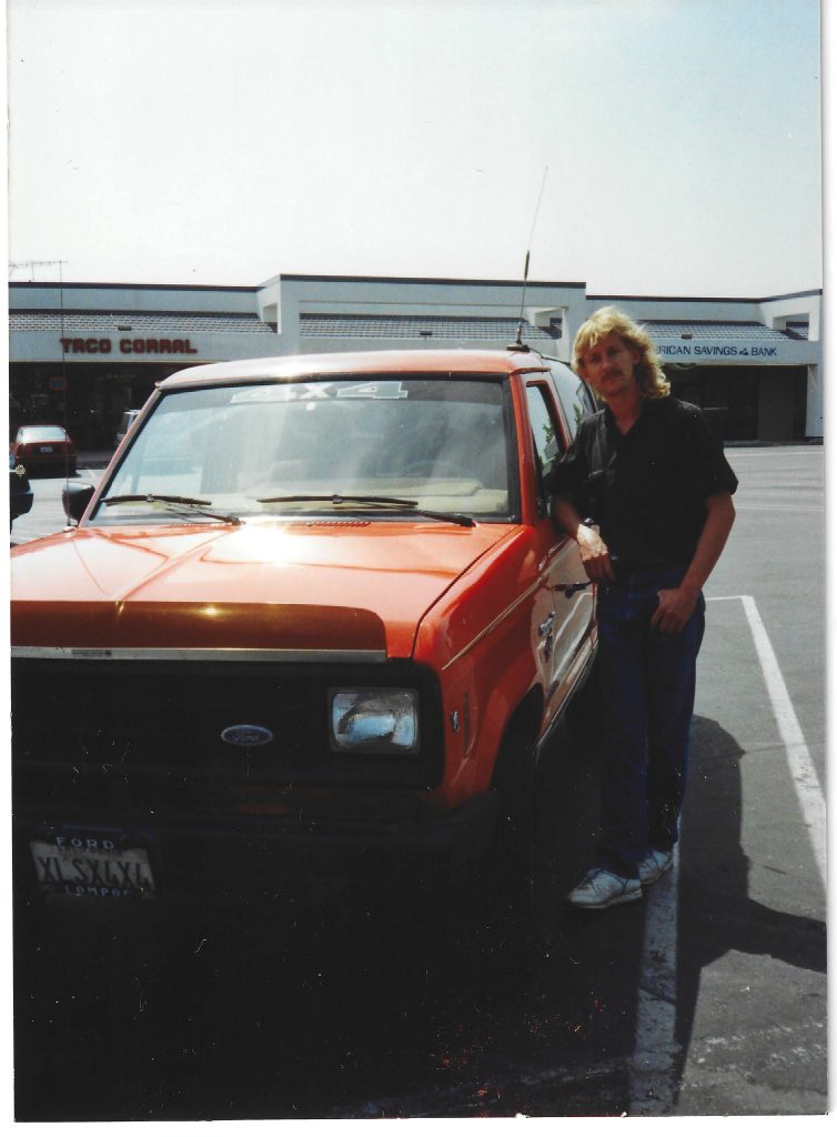 Man standing next to an orange Ford truck in a parking lot, with Taco Corral and American Savings Bank visible in the background, showcasing classic vehicle detailing and restoration potential.